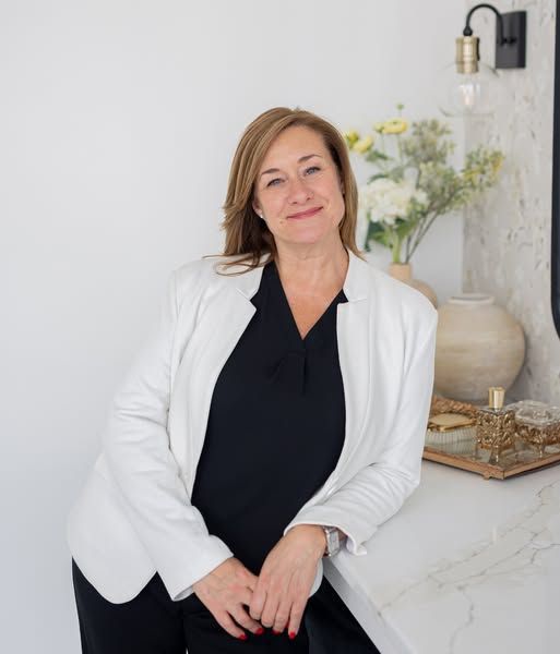 Woman in white blazer leans on counter, smiling.