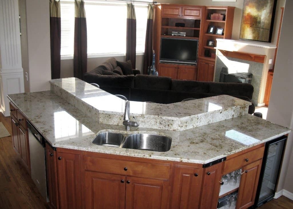 A granite countertop island with a sink in a kitchen, overlooking a living room with a TV and fireplace.