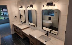 Bathroom with three sinks, mirrors, and light fixtures. White countertops and wood cabinets. Dark tiled floor and white plank walls.