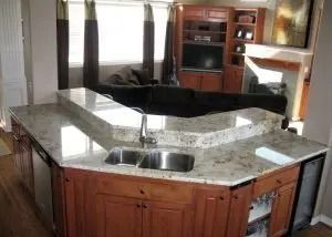 Kitchen island with granite countertop, sink, and cabinets; a living room is in the background.