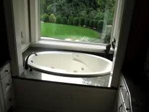 Oval bathtub set into a granite counter, under a window with a view of a green lawn and trees.