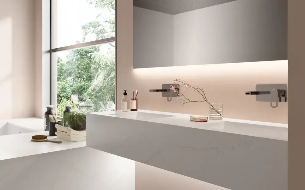 Modern bathroom with white countertop, large mirror, and wall-mounted faucets, next to a window with greenery.