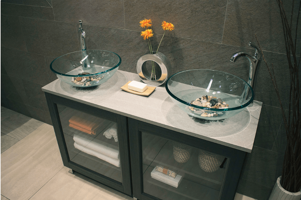 Bathroom vanity with two glass bowl sinks, gray cabinets, towels, and decorative flowers.