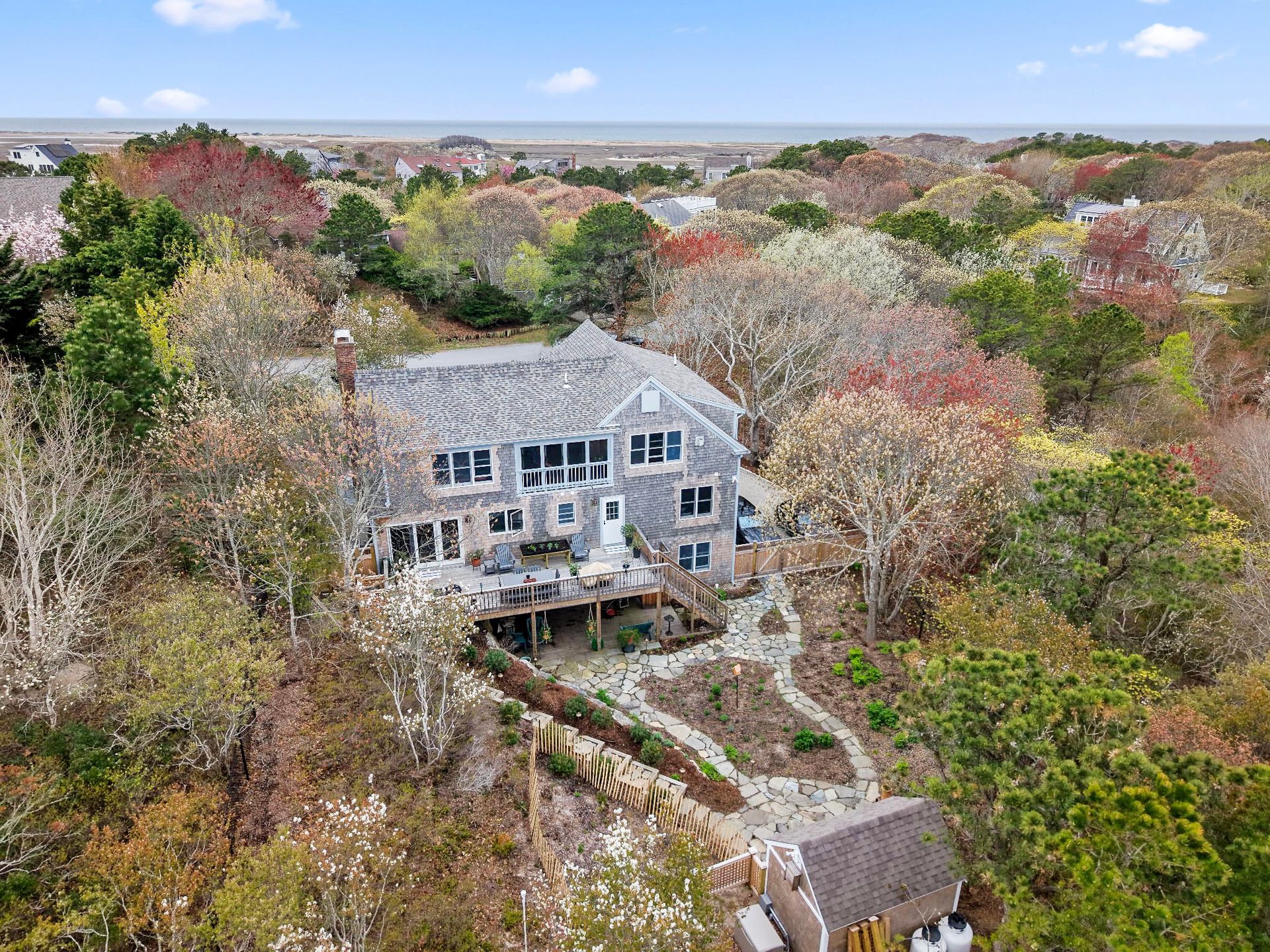 Coastal house with gray shingles and wooden decks, surrounded by trees, ocean view in the distance.