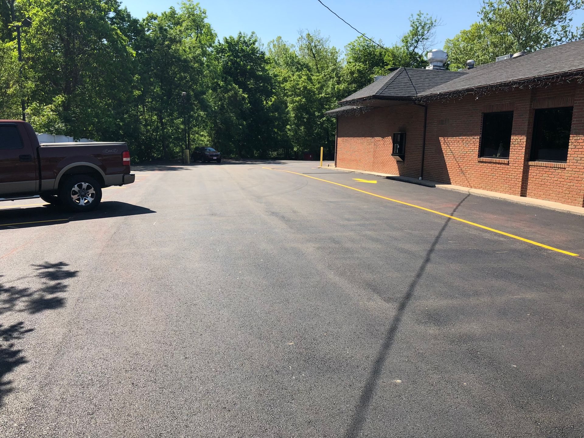 Empty parking lot beside a brick building, with a pickup truck and trees in sunlight.
