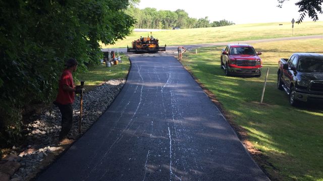 Freshly paved driveway through grassy field, with a worker, parked cars, and a paver in the distance