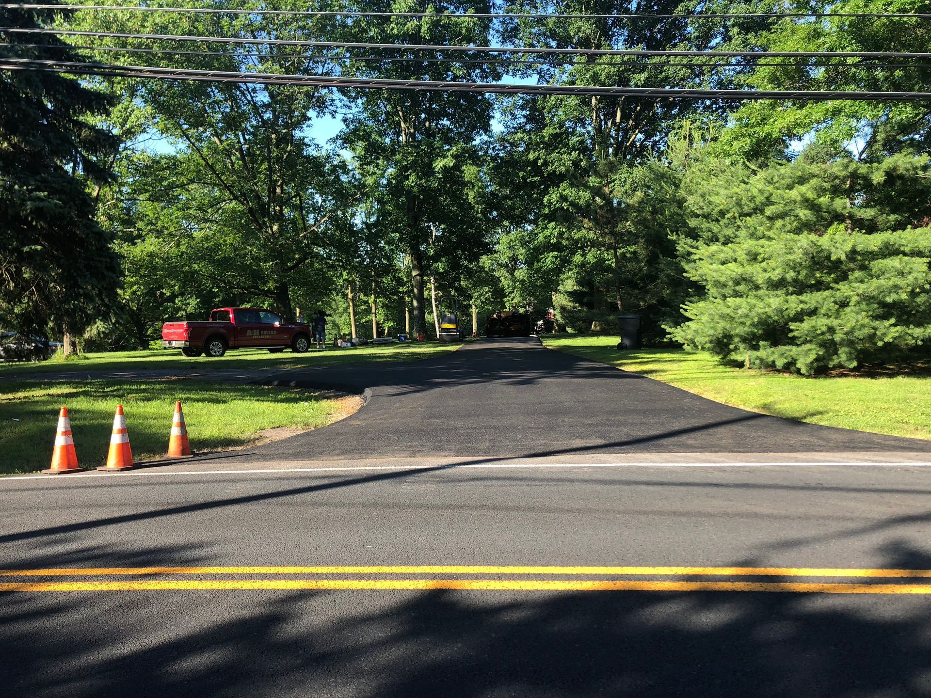 Tree-lined suburban road blocked by orange cones, with a dark driveway and yellow center lines