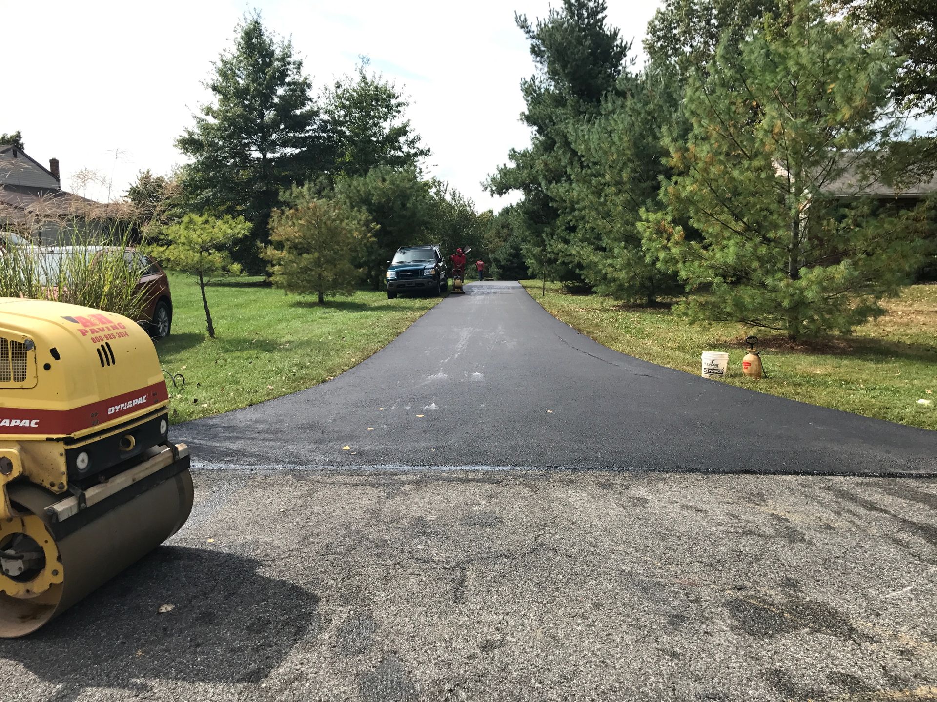 Freshly paved driveway leading to a house, with a roller on the left and trees lining both sides.