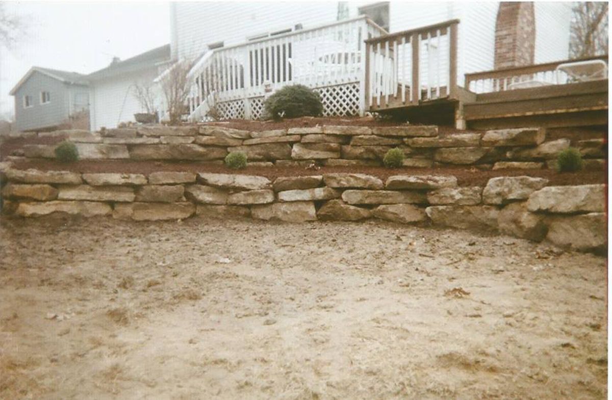A stone wall is in front of a house with a deck.