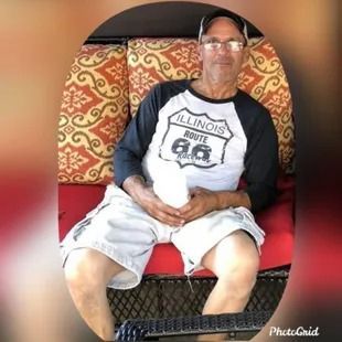 Man sitting outdoors, wearing Route 66 shirt and shorts, holding a white object.