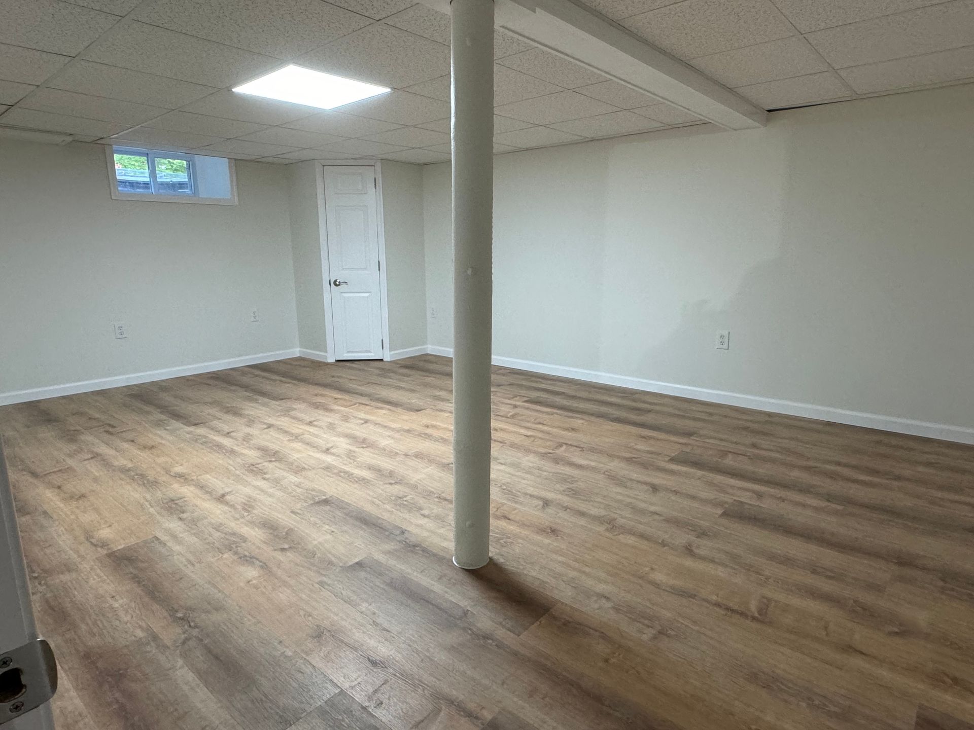 Empty, finished basement with wood-look flooring, white walls, support column, and door.