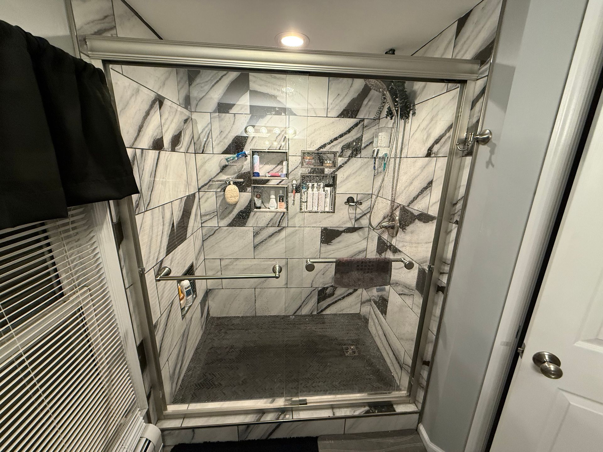 Shower with marble-patterned tiles, glass door, and silver fixtures. Includes a shelf for toiletries and a grab bar.