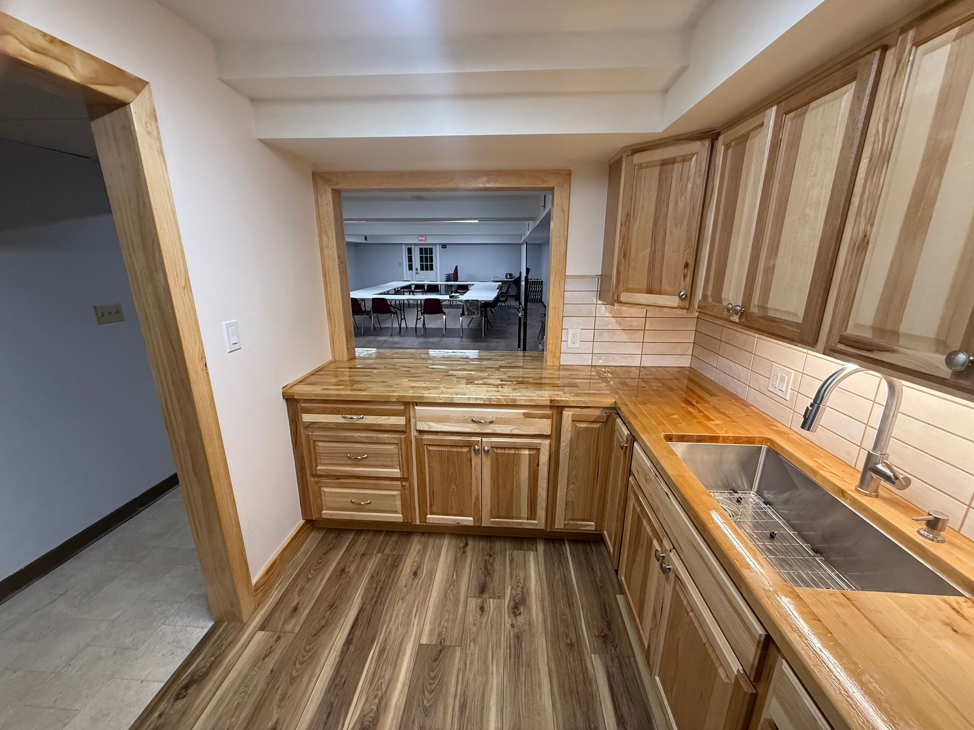 Kitchen with wooden cabinets, countertop, and sink, overlooking a room with tables and chairs.