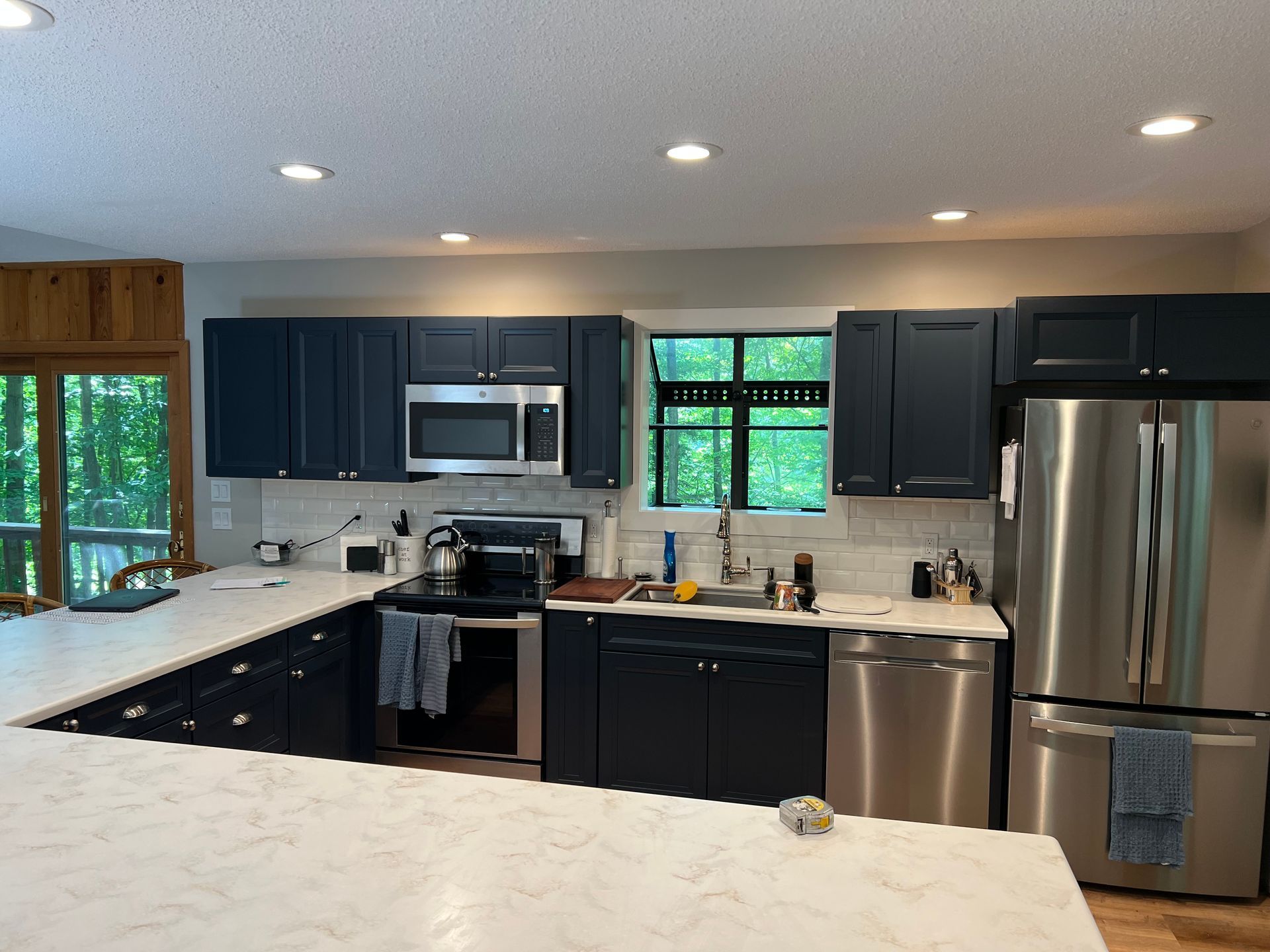 Navy blue kitchen with stainless steel appliances, white countertops, and a window overlooking greenery.
