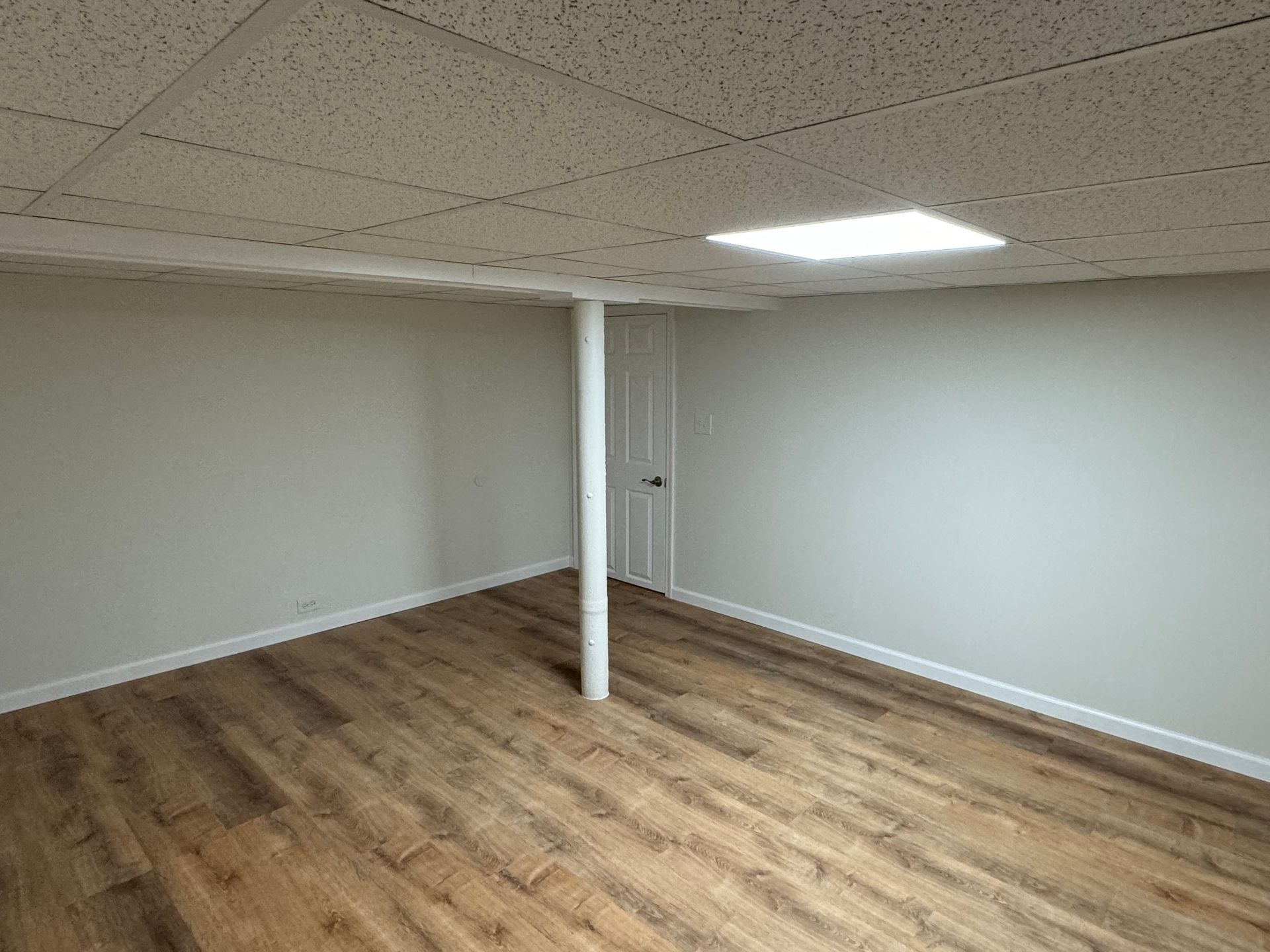 Empty room with light wood-look floor, white walls, and a dropped ceiling with a fluorescent light. A support pole is in the center.