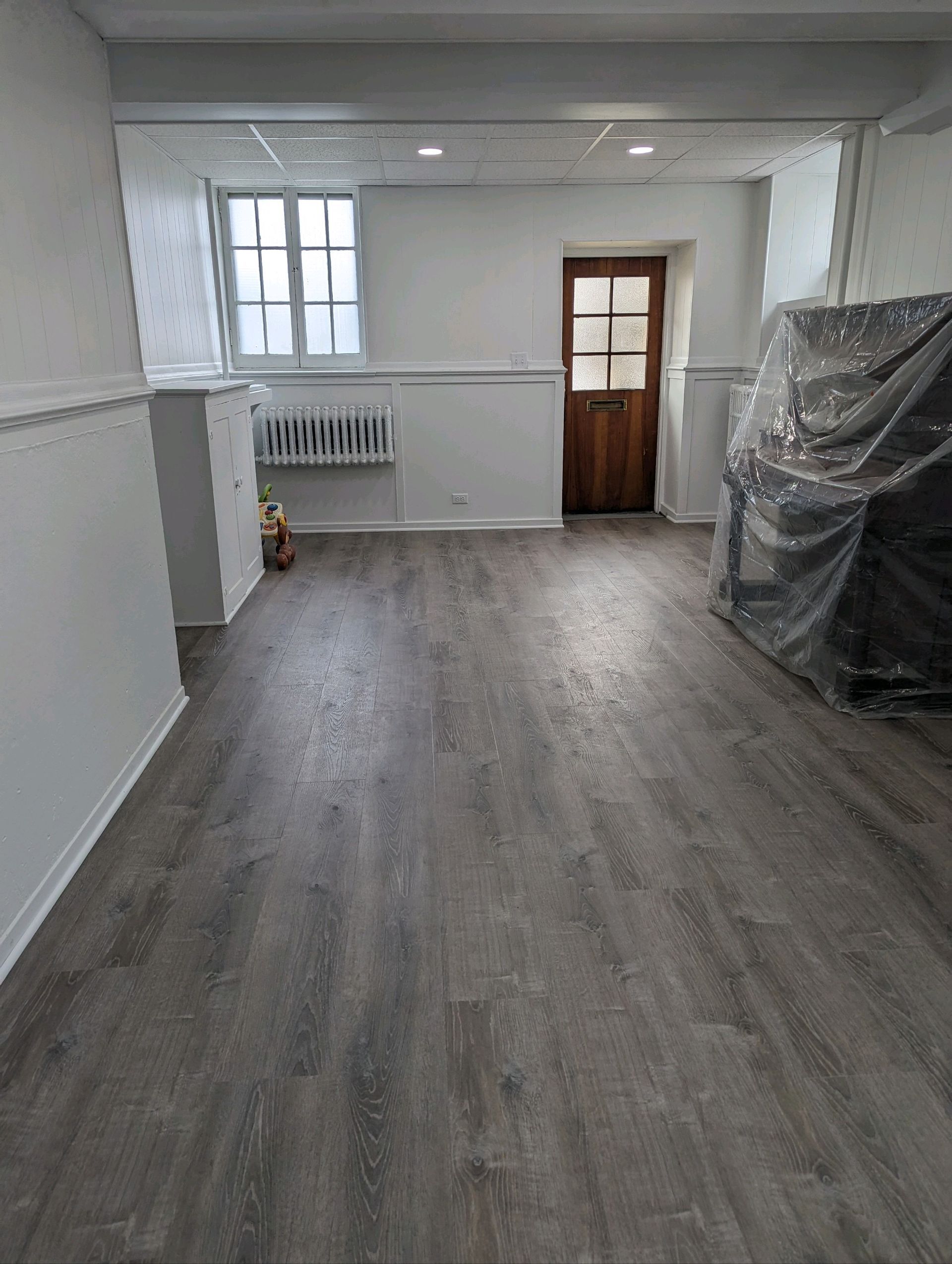 Empty room with gray wood-look flooring, white walls, and a wooden door. A window is in the back.