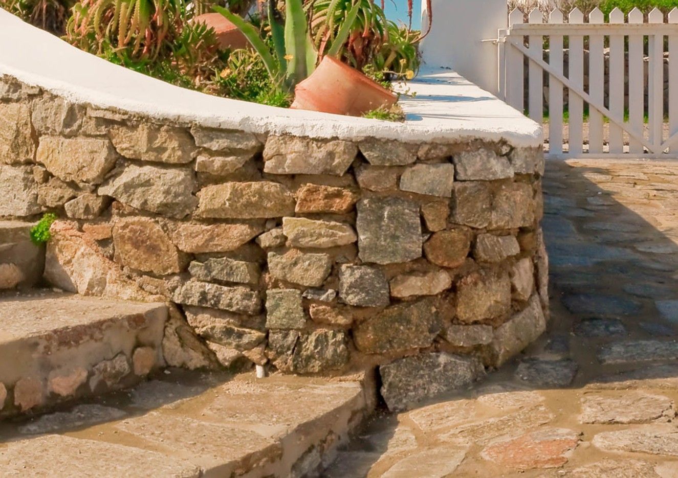 A stone retaining wall curves along a cobblestone patio near a white wooden gate and some green plants.