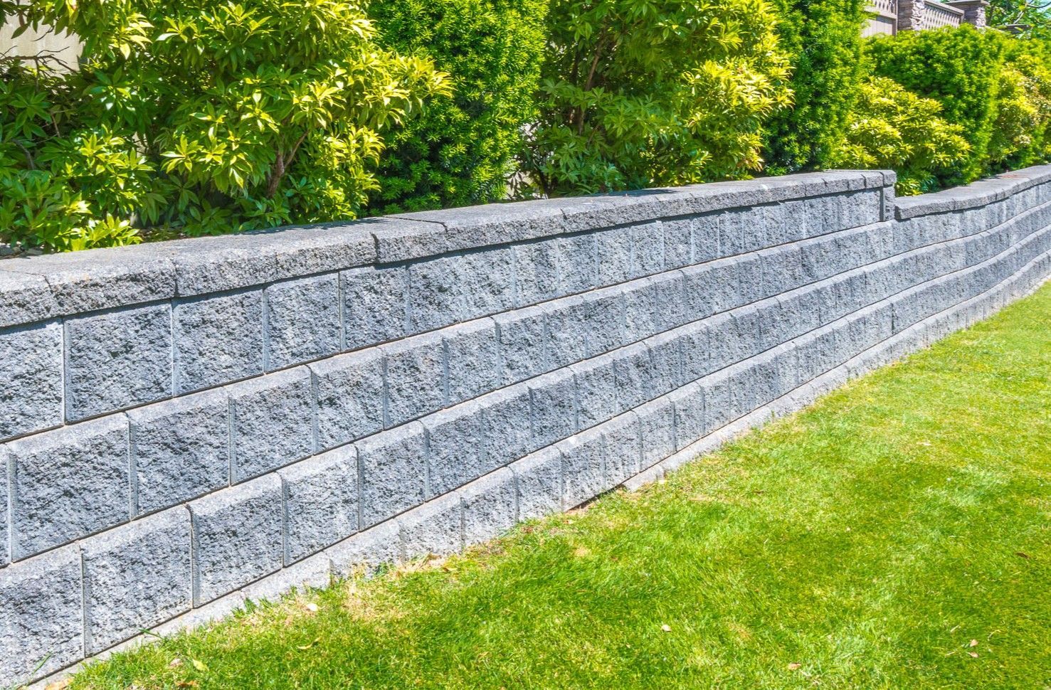 A grey, textured retaining wall made of stacked concrete blocks, bordering a green lawn with shrubs in the background.