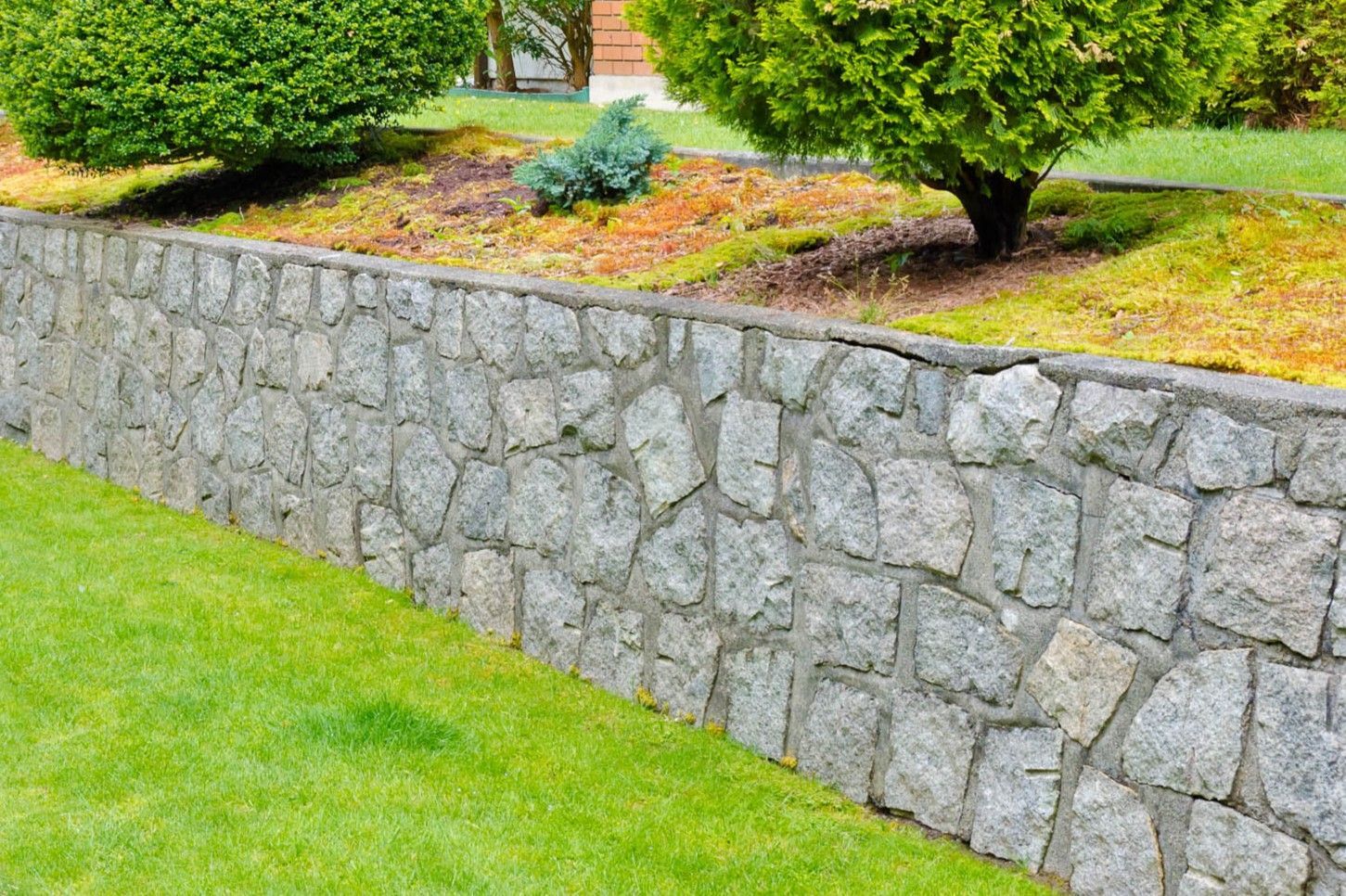 A stone retaining wall borders a green lawn with landscaped shrubs and trees in the background.