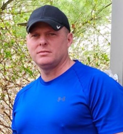 Man in a black cap and bright blue shirt standing outdoors by greenery
