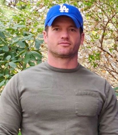 Man in a blue LA cap and olive shirt standing outdoors in front of greenery.