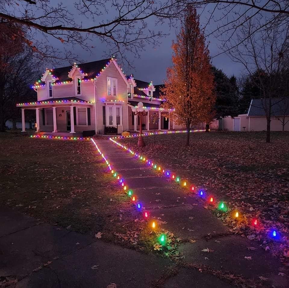 Victorian house at dusk outlined with colorful Christmas lights, with two lit paths leading to the porch