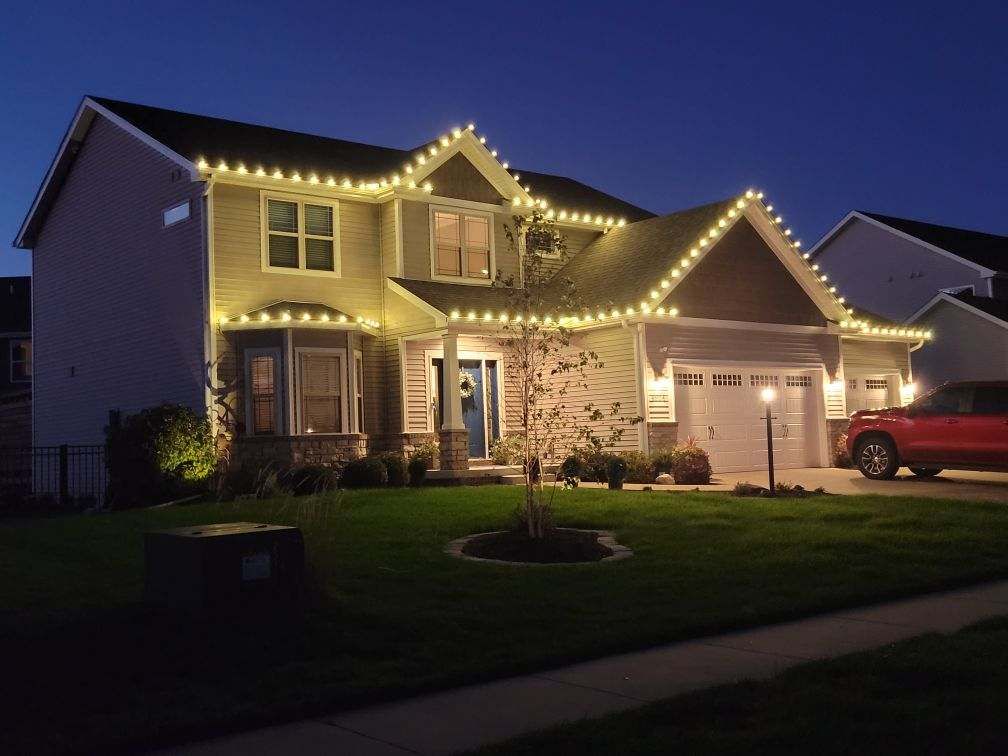 Two-story suburban house outlined with warm holiday lights at dusk, with a red truck in the driveway