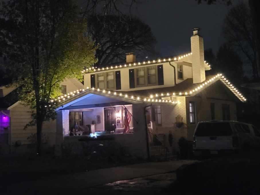 House at night outlined with warm white holiday lights, with a lit porch and parked vehicle in front.
