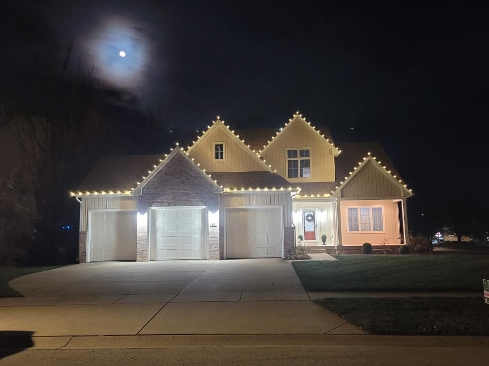 Two-story house at night with lit holiday roof lights, three garage doors, and a full moon overhead