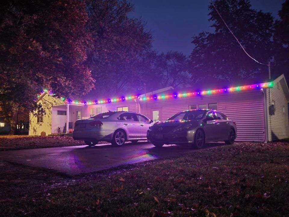 Two cars parked outside a house lit with colorful string lights at night