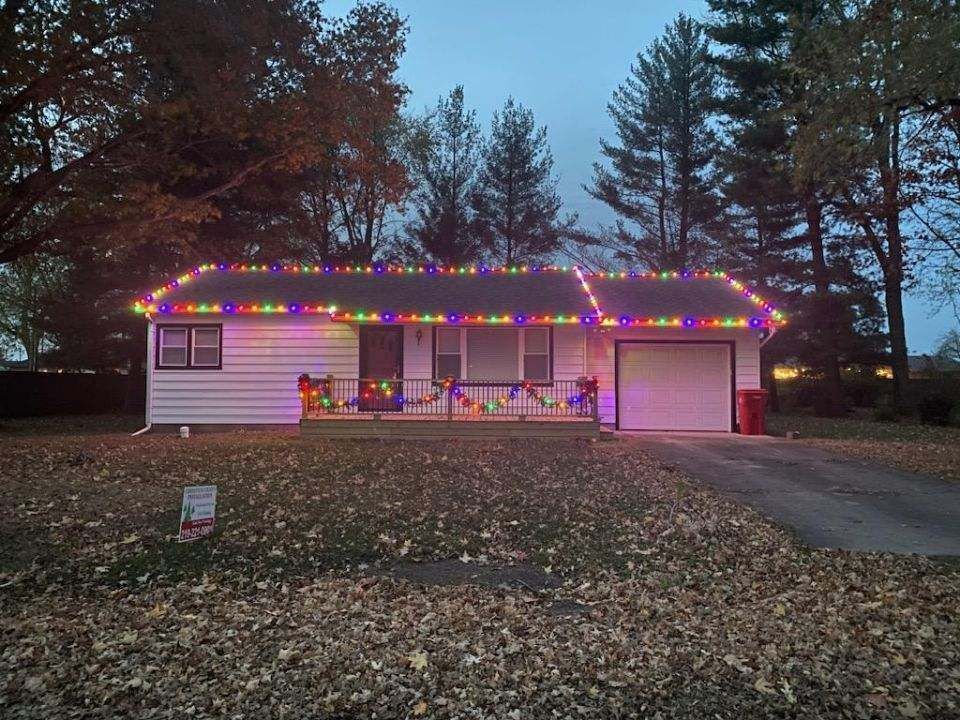 House decorated with colorful Christmas lights at dusk, with trees and a driveway in front.