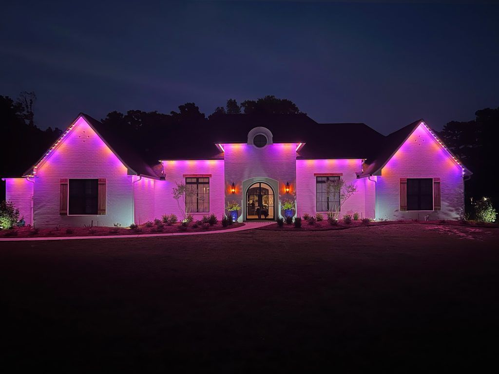 House lit with pink-purple holiday lights at night, with a dark yard and dark sky.