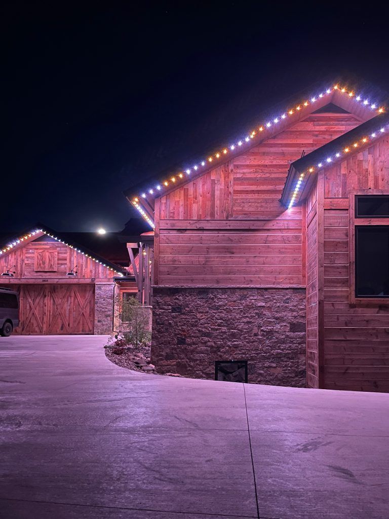 Snowy driveway beside a wooden house and garage at night, lit by purple-white roof lights