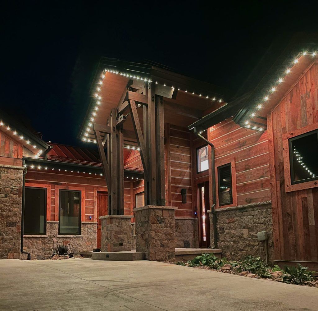 Rustic lodge entrance at night, with red wood siding, stone pillars, and string lights glowing along the roof.