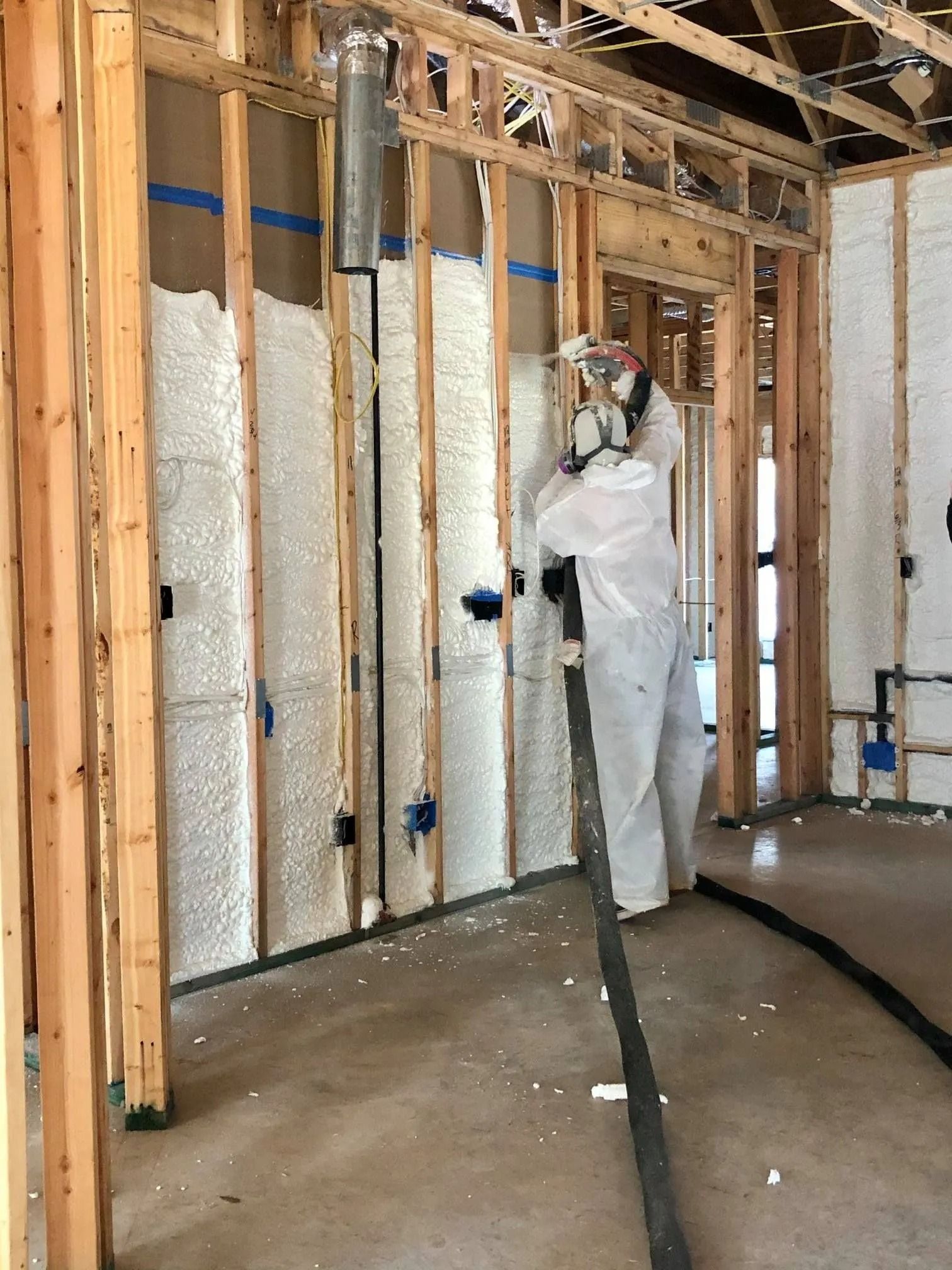 Person in protective suit spraying foam insulation into a wooden wall frame in a building.