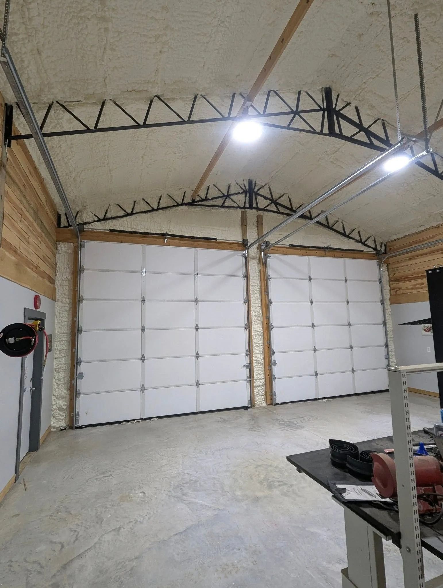 Interior view of a workshop with two large white garage doors, spray foam insulation, and metal trusses.