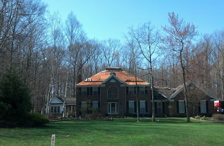 A two-story brick house with a partially exposed, reddish-brown roof deck sits in a wooded yard on a sunny day.