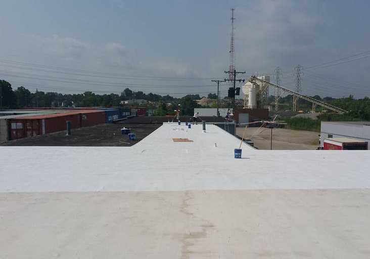 A white flat roof surface leading to an industrial area with power lines, silos, and shipping containers under a blue sky.