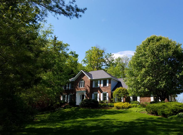 A two-story brick house with white shutters sits on a lawn surrounded by mature trees under a clear blue sky.