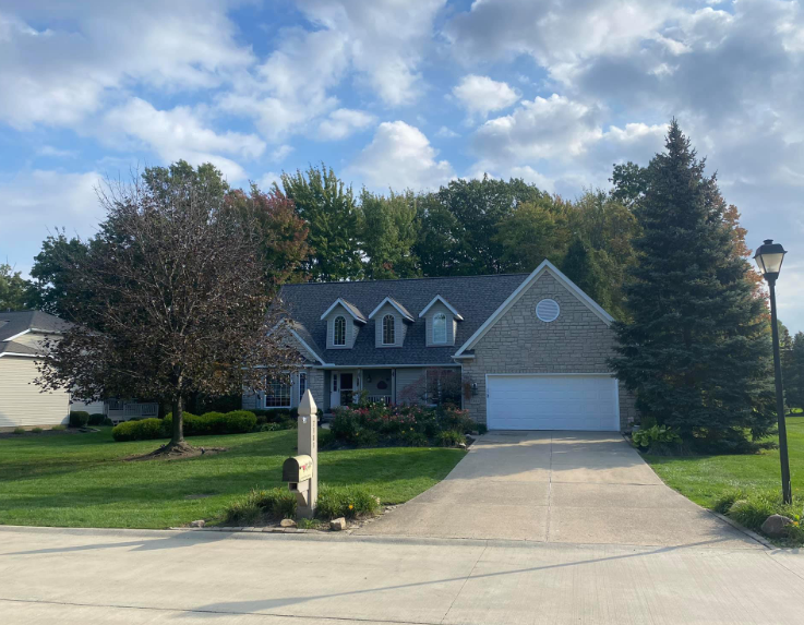 A single-story brick house with a two-car garage, dormer windows, and a lush green front lawn under a partly cloudy sky.
