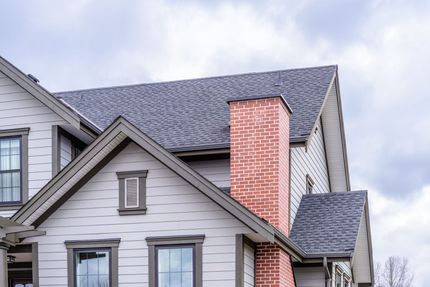 A section of a house exterior featuring dark shingles, light gray siding with dashed accents, and a red brick chimney.