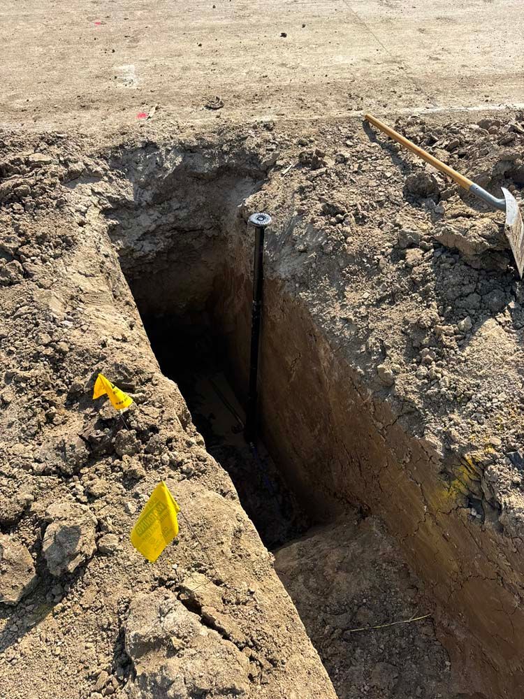 A deep trench in dirt with a metal pole and shovel visible. Yellow flags mark the edges.