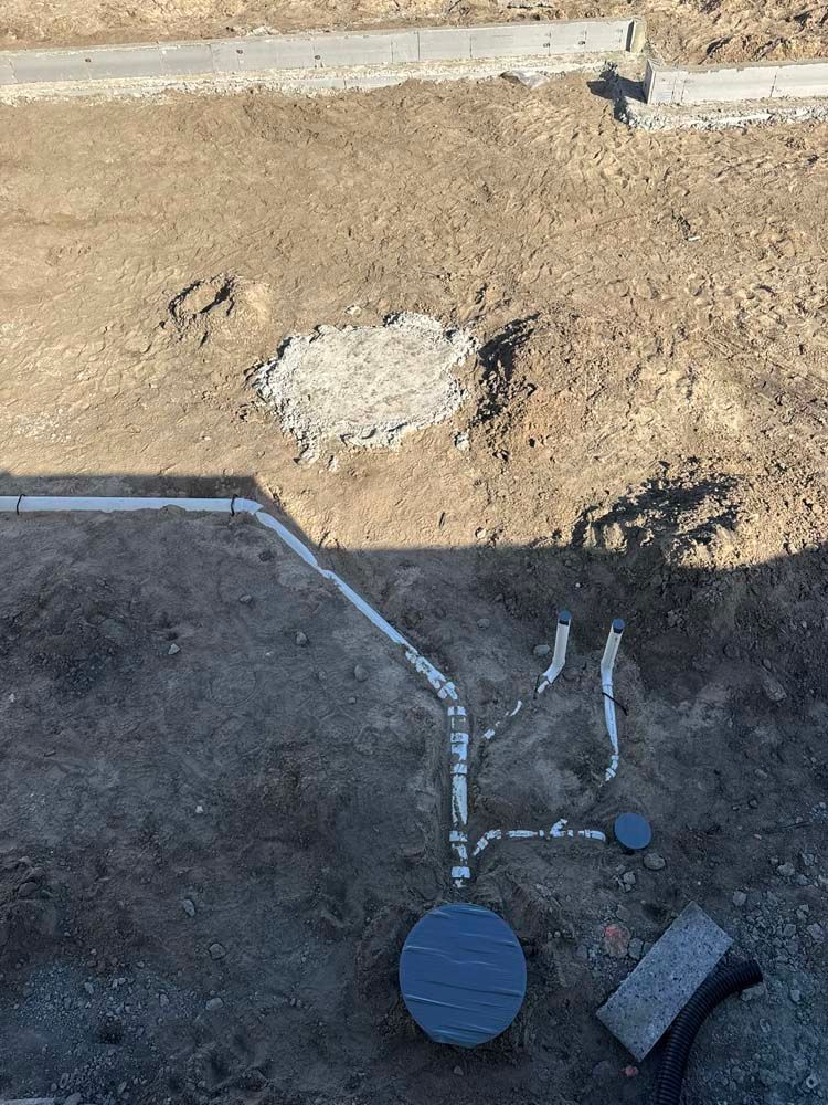Pipes and a gray circular object are exposed in a dirt area, with a concrete section.