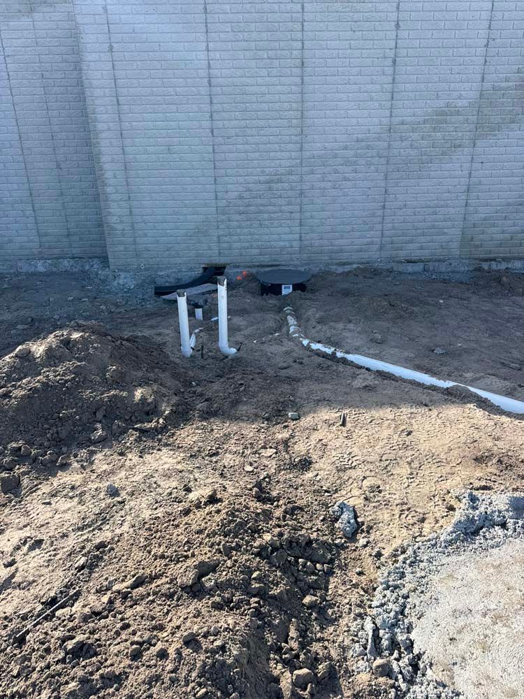 Pipes protruding from the ground against a gray brick wall, in a dirt setting.
