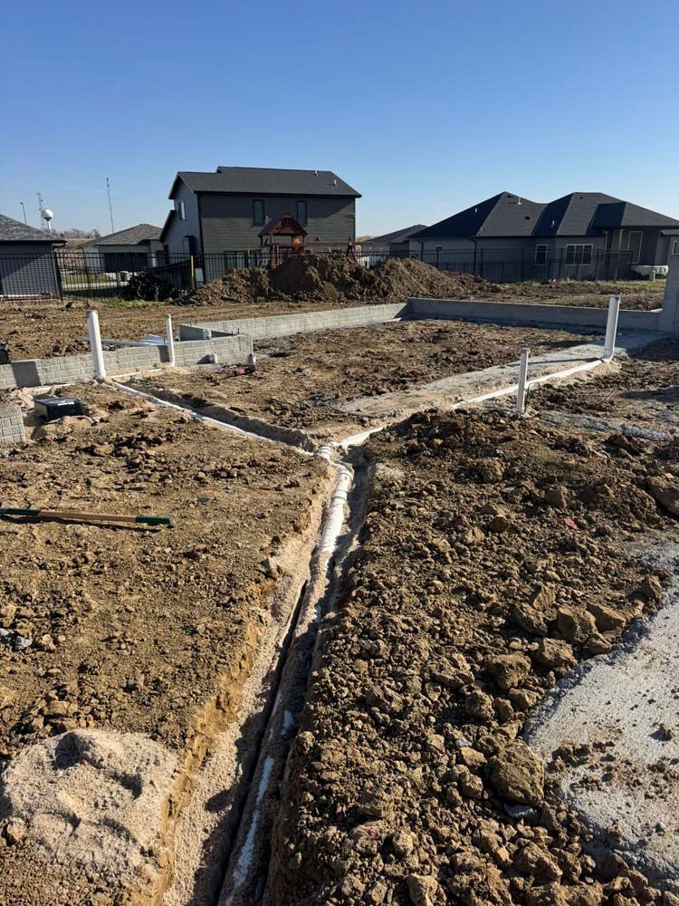 Construction site with trenches for plumbing pipes, foundations, and houses in the background. Sunny day.