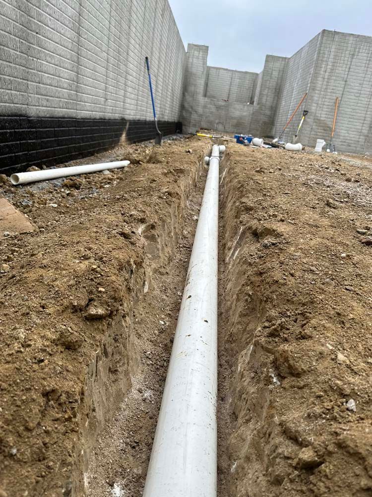 White PVC pipe laid in a trench, near a brick wall under construction.
