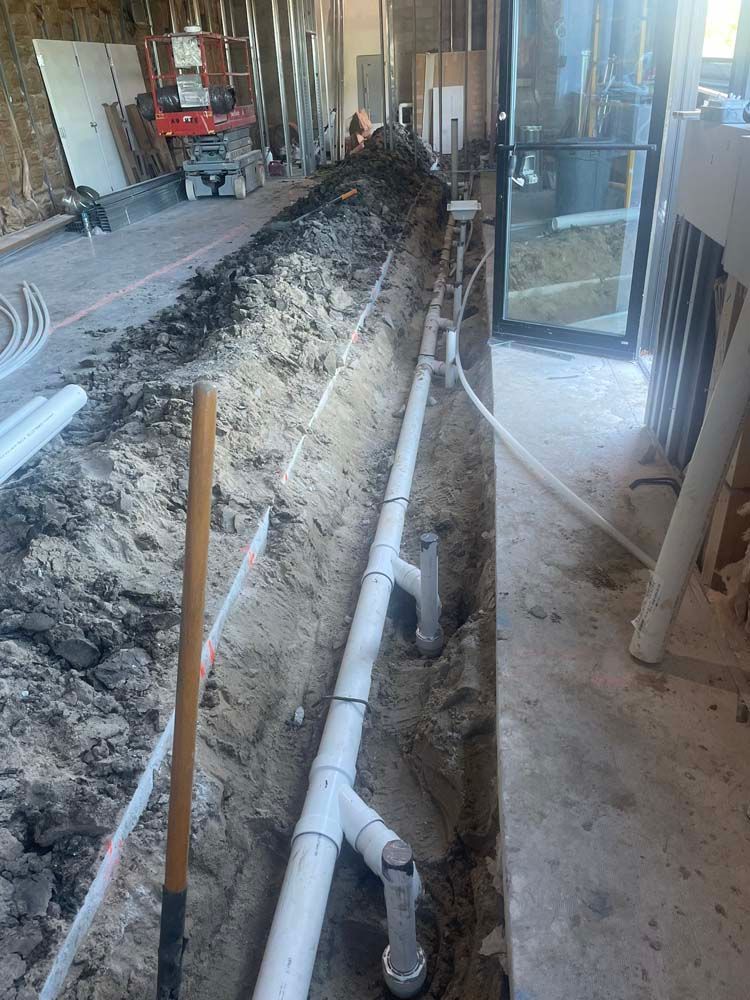 Trench with white pipes and connections; construction site, shovel in foreground, glass door on right.