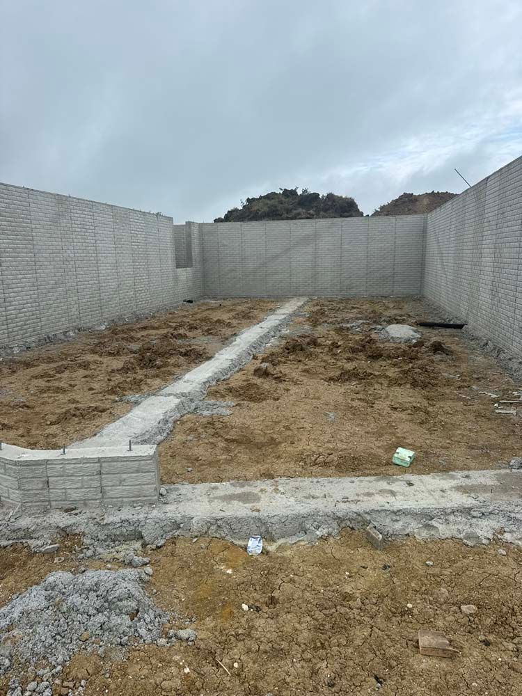 Construction site: grey brick walls enclose a dirt lot. A partially built walkway of bricks is visible. Cloudy sky.