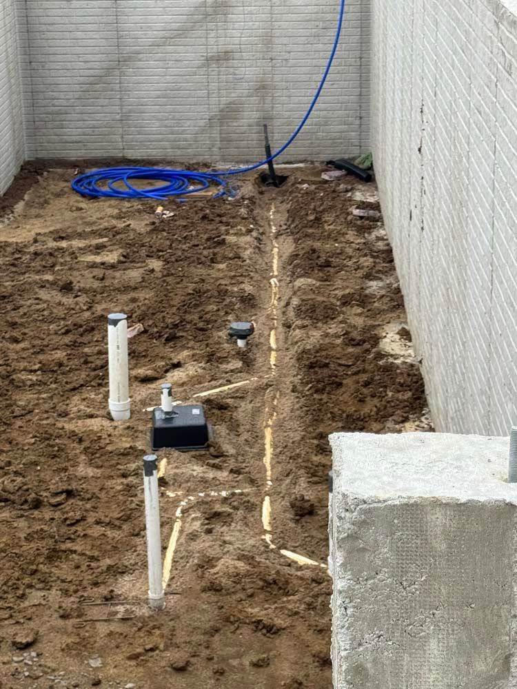 Dirt area with trenches, pipes, and a blue hose against concrete walls.