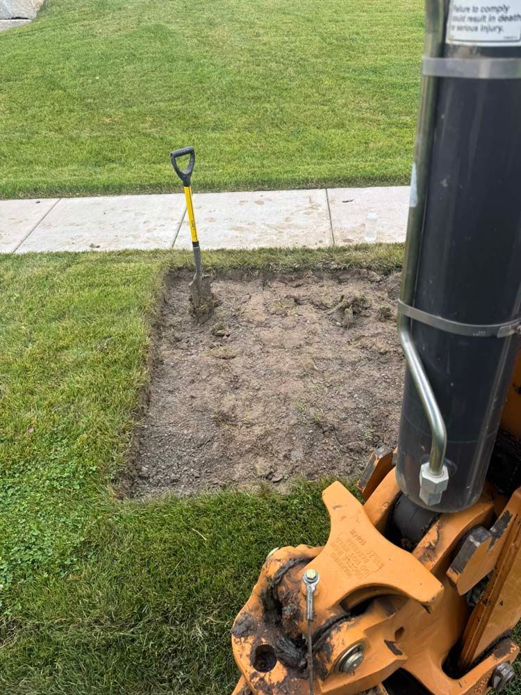 A small square hole dug in a grassy lawn next to a sidewalk with a shovel in the hole, and a machine nearby.